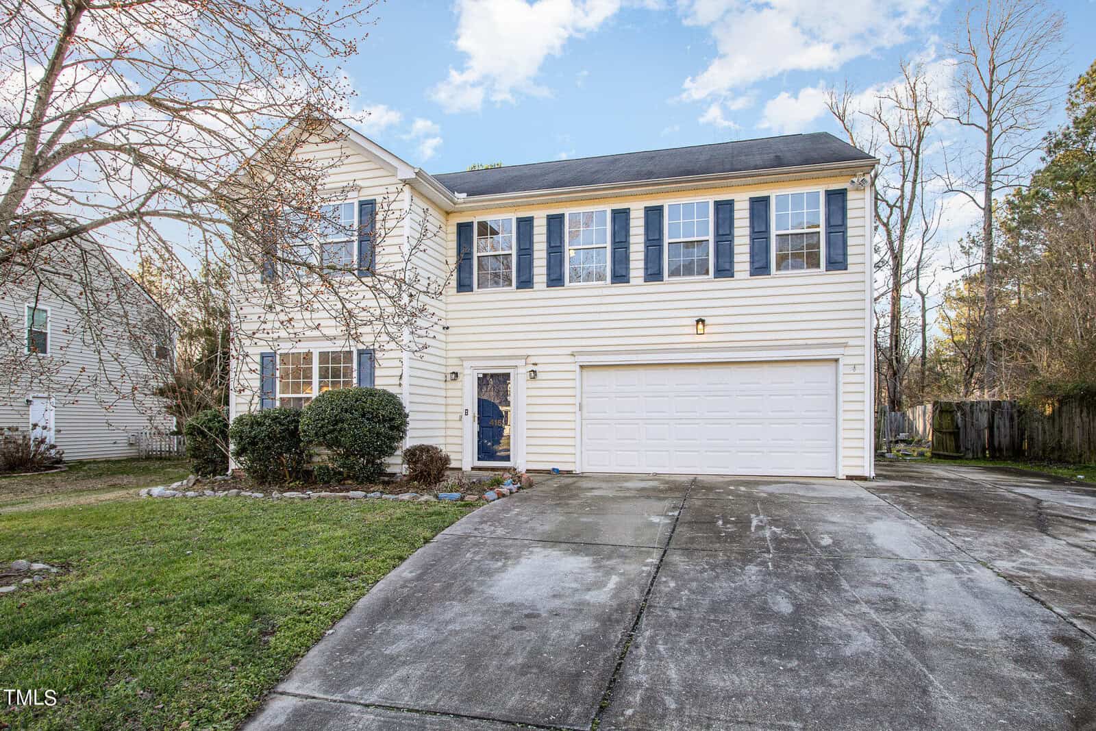 The two-story house at 416 Mayview Drive in Creedmoor boasts light siding and dark shutters beneath a blue sky. It features a two-car garage and a curved driveway, with well-maintained grass and bushes in the front yard, while bare trees surround the property.