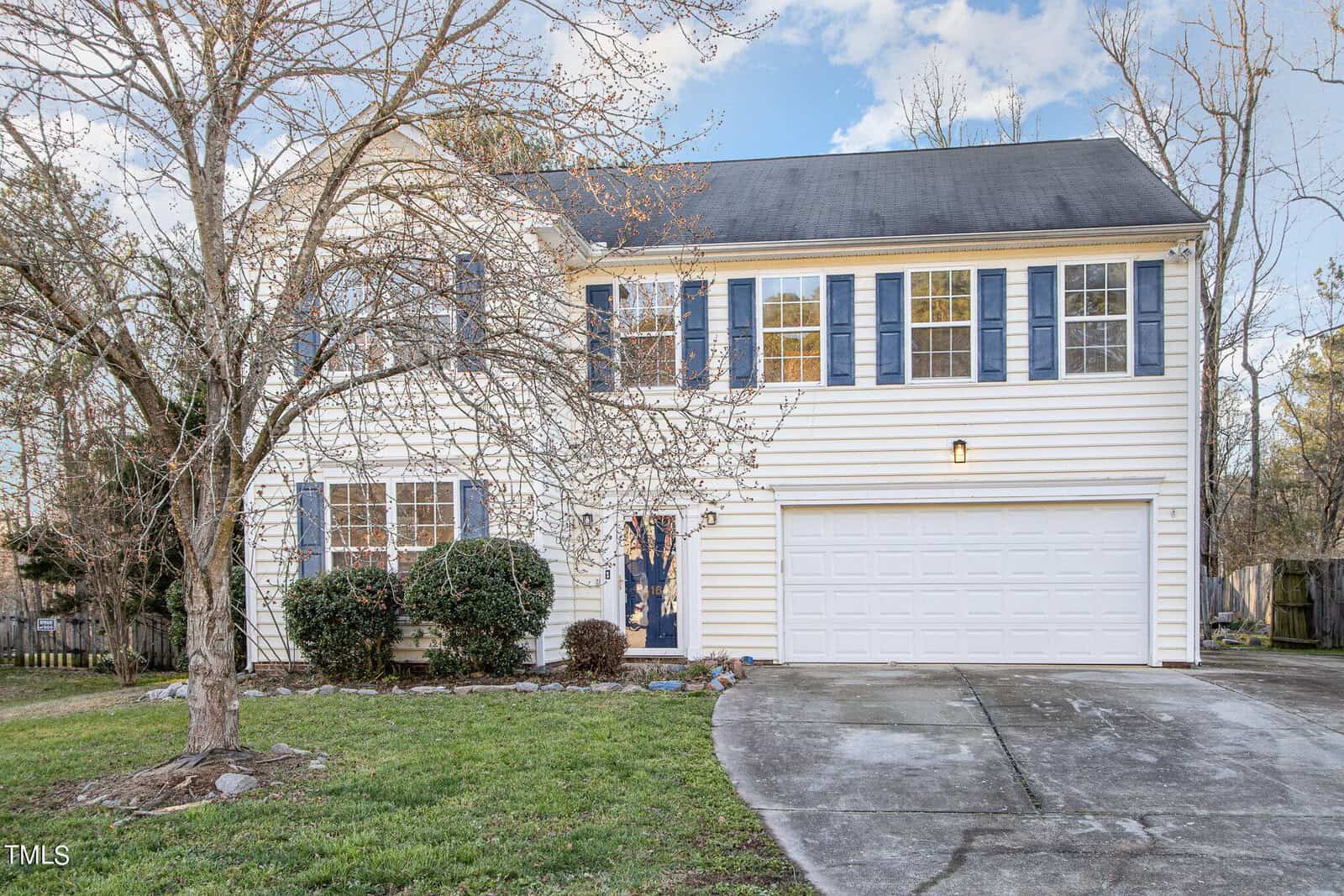 A two-story white house with blue shutters and a two-car garage. It features a driveway, bare trees, and shrubs in the front yard. The sky is clear.
