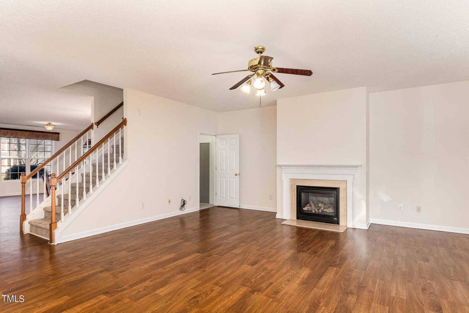 A spacious, empty living room with hardwood floors and a ceiling fan. There is a fireplace with a white mantel on the right wall. A staircase with a wooden railing leads upstairs, and an open doorway leads to another room.