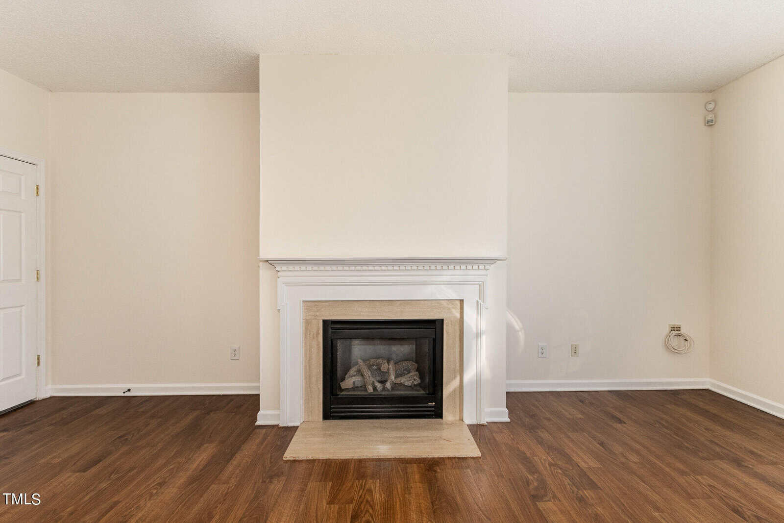 A cozy room with beige walls features a centered fireplace framed by white trim. The floor is dark wood, and there is a closed door to the left and an electrical outlet on the right wall.