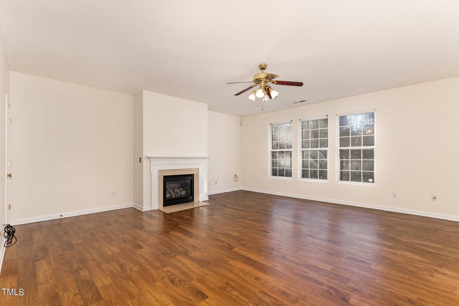 Empty living room with wooden floors, white walls, and large windows. A white fireplace is centered against one wall, and a ceiling fan with lights is overhead. Natural light brightens the space.
