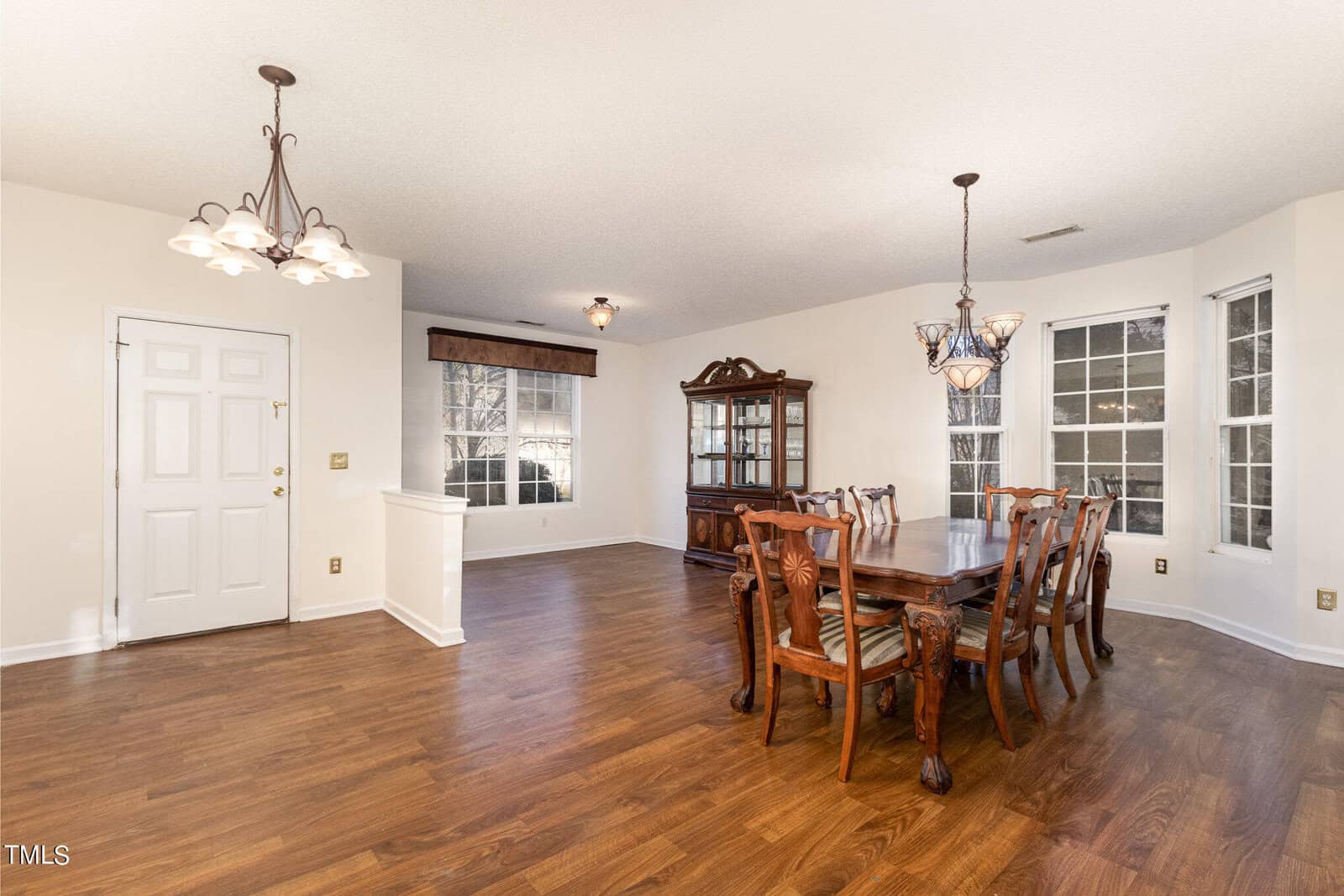 A dining room with hardwood floors, a rectangular wooden table, and six chairs. Two chandeliers hang from the ceiling. Large windows allow natural light in, and a wooden cabinet is against the wall. There's a white door on the left.