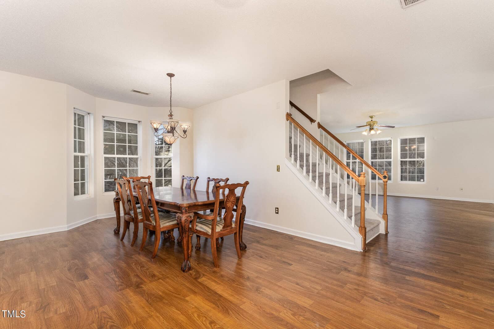 A dining area with a wooden table and chairs next to a staircase leading upstairs. The room has hardwood floors and large windows, offering natural light. A chandelier hangs above the table, and a ceiling fan is visible in the adjacent room.