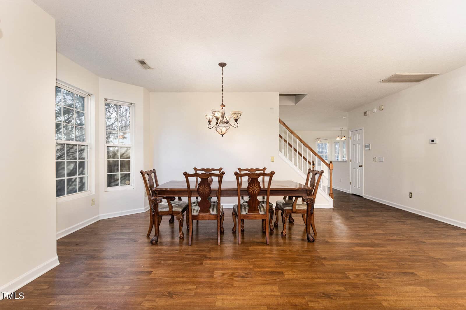 Dining area with a wooden table and six chairs. A decorative chandelier hangs above the table. The room features large windows on the left and hardwood flooring. A staircase and doorways are visible in the background. Bright, neutral walls.