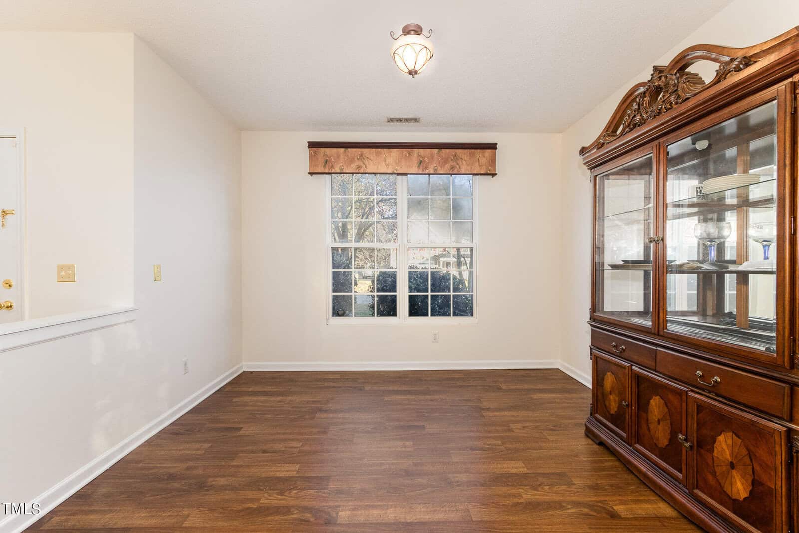 An empty room with hardwood floors and a large window. A decorative china cabinet with glass doors stands on the right. The walls are white, and a small ceiling light fixture provides illumination.