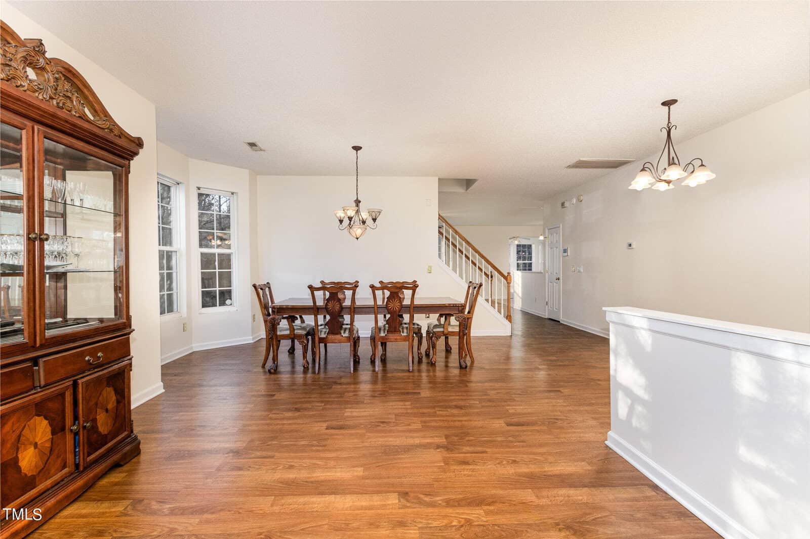 A bright dining area with wooden floors, a chandelier, and a wooden dining table set for six. A wooden china cabinet is on the left, and a white staircase with wooden railings leads upward in the background. Natural light filters through large windows.