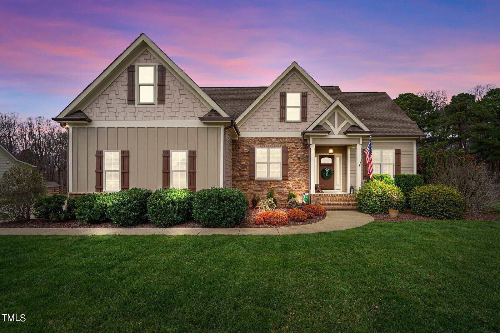A charming house on 3853 Ironwood Drive with a mix of stone and siding under a twilight sky. It proudly displays an American flag by the entrance, while the well-maintained yard in Franklinton features lush bushes and a vibrant lawn against a sky painted in purple and pink hues.