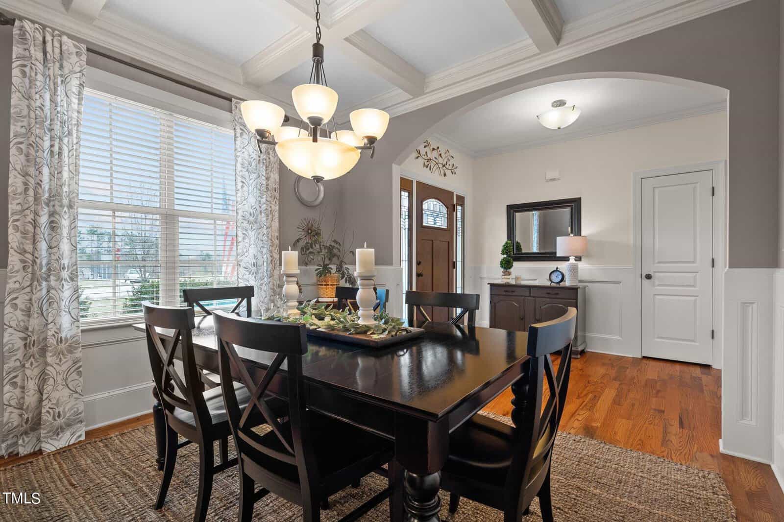 A dining room with a dark wooden table and six chairs. The table has three candles and decorative items. A modern chandelier hangs above. Light streams in through a window with patterned curtains. The room has hardwood floors and an entryway in the background.