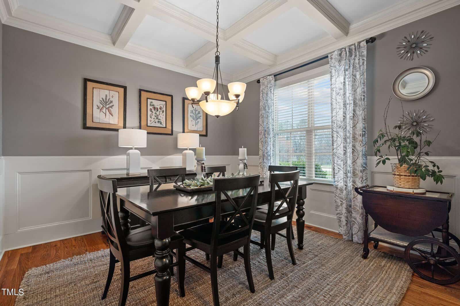 A dining room with a dark wood table and six chairs, a chandelier above, and a sideboard with two lamps. The walls are gray with white wainscoting, decorated with framed art. A window with patterned curtains and natural light brightens the space.