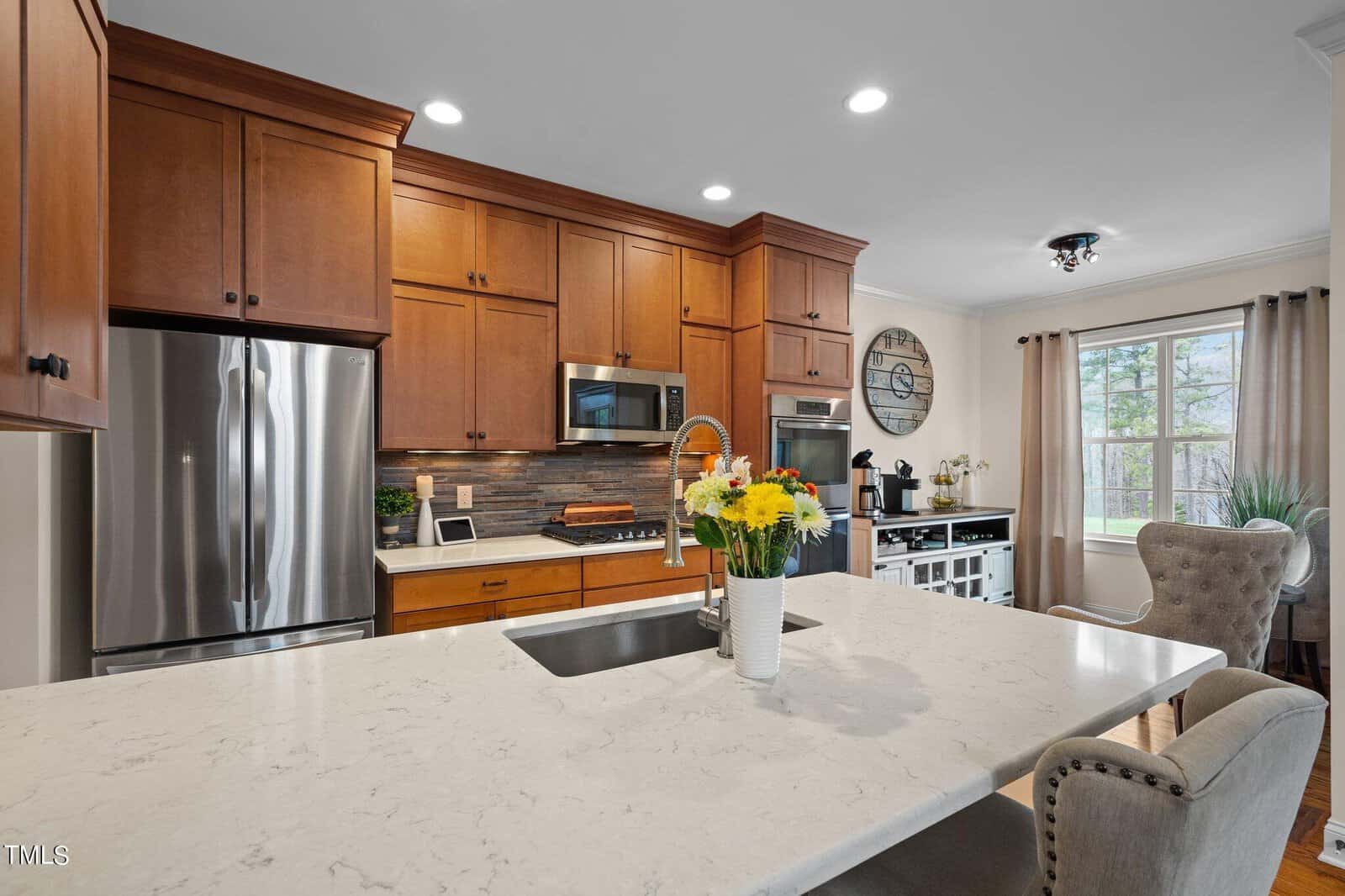 A modern kitchen with wooden cabinets, stainless steel appliances, and a marble island with a sink. A vase with colorful flowers sits on the island. There's a large window with curtains and a cozy chair nearby.