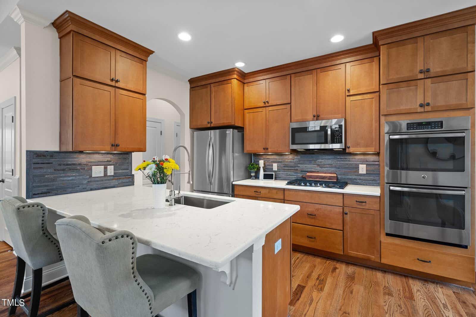 A modern kitchen with wooden cabinets, stainless steel appliances, and a marble island with two cushioned chairs. The backsplash is gray with a brick pattern, and a vase with flowers decorates the island. Warm lighting brightens the space.