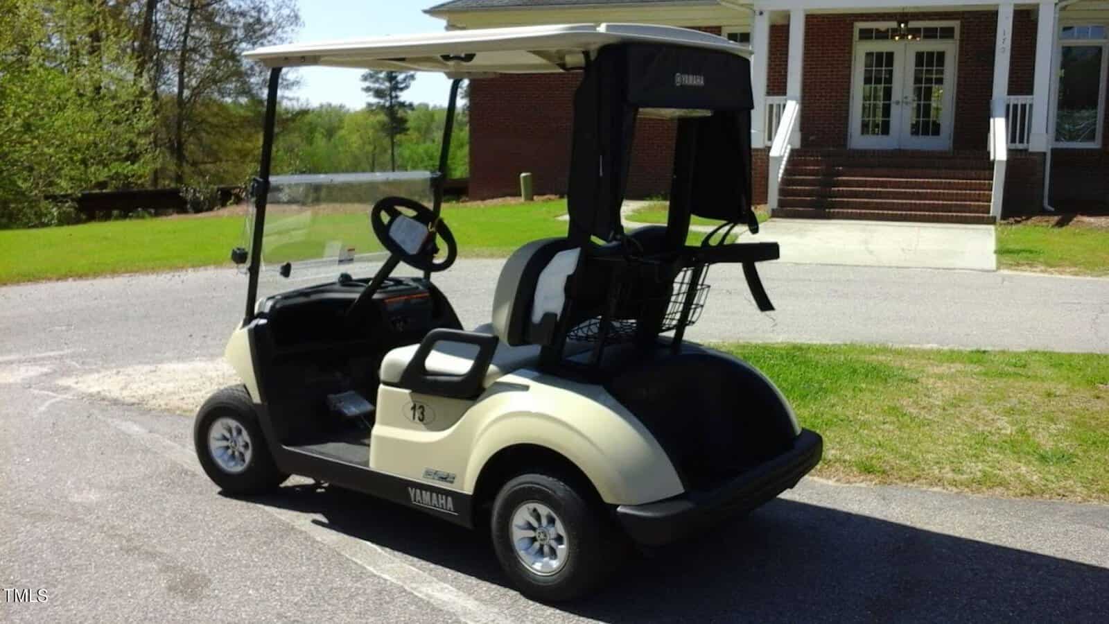 A beige Yamaha golf cart parked on a paved driveway in front of a brick building with white columns and steps, surrounded by greenery and trees.