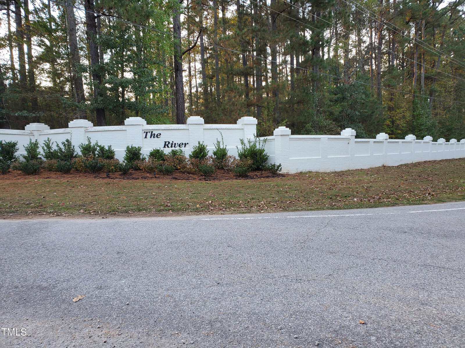 A white brick wall with shrubbery in front and the words "The River" displayed, bordered by a forest of tall pine trees and a paved road in the foreground.