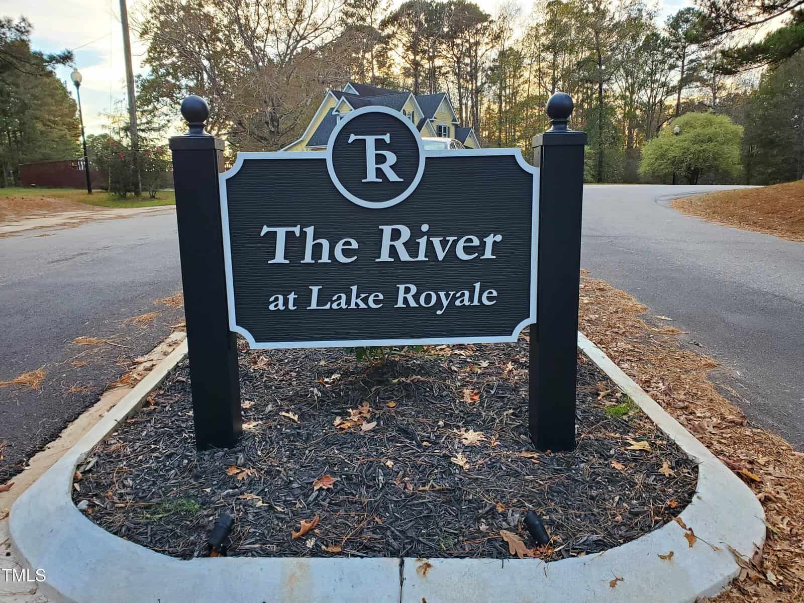 A black and white sign reads "The River at Lake Royale" on a mulched area by a road, with trees and houses in the background.