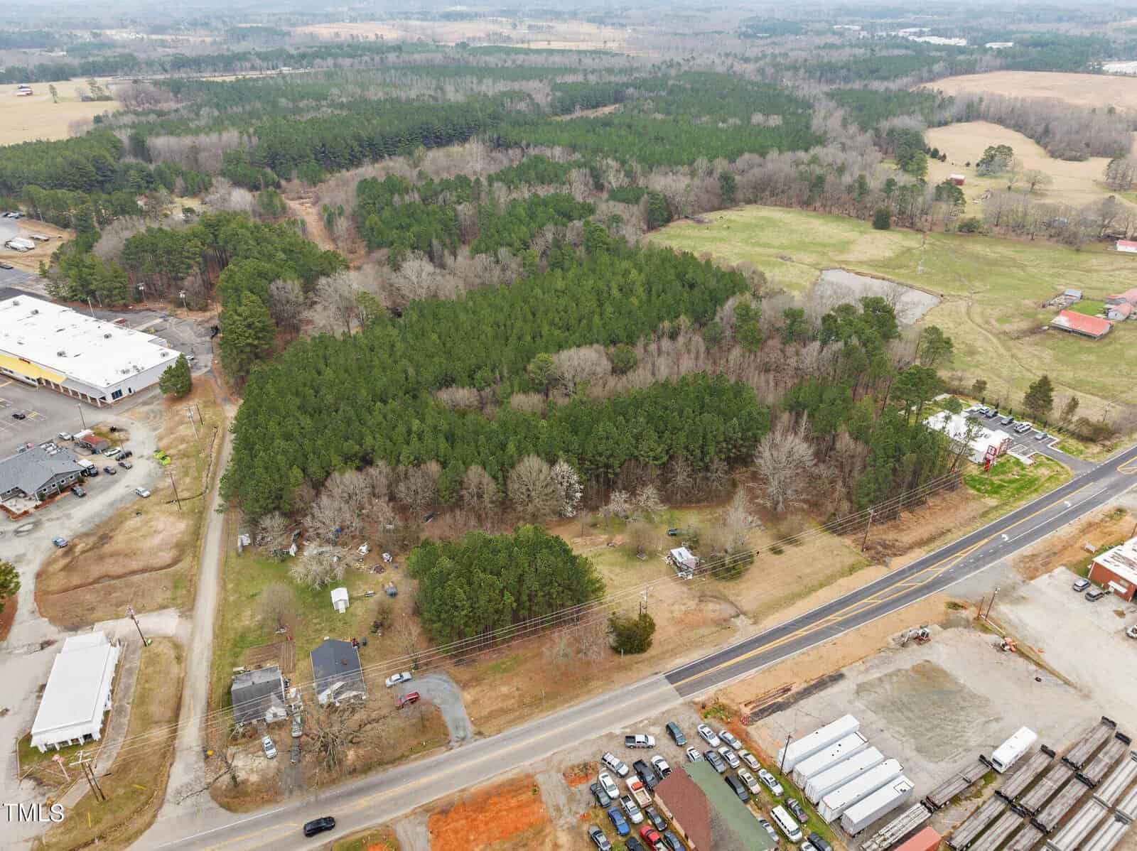 Aerial view of a rural landscape with a dense forest in the center. Surrounding the forest are roads, scattered houses, a large warehouse, open fields, and a few vehicles. The background features more forested areas and distant fields.