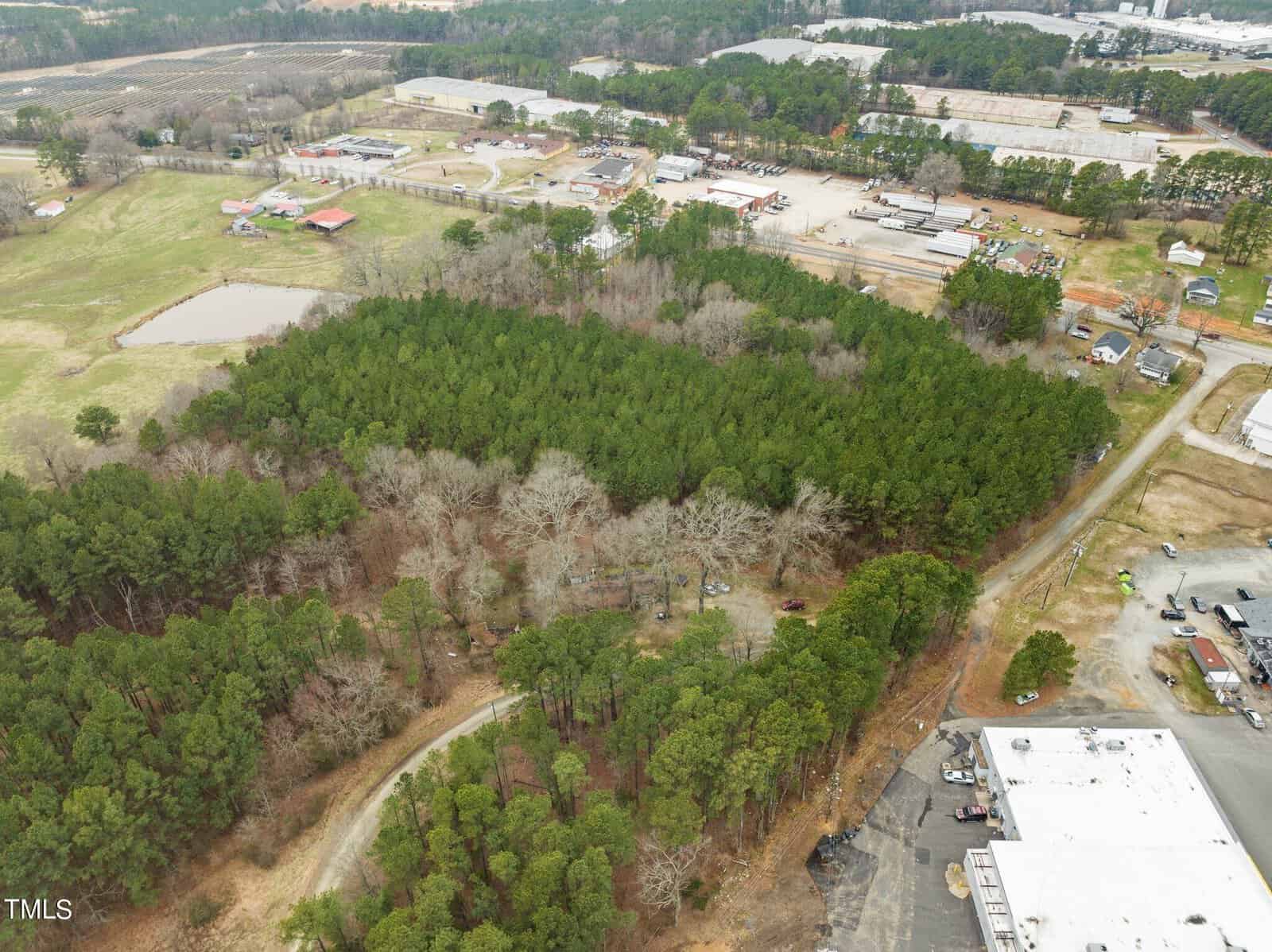 Aerial view of a wooded area with dense trees surrounded by industrial buildings and roads. The landscape includes grassy fields and a small pond, with various structures and vehicles visible in the surrounding area.