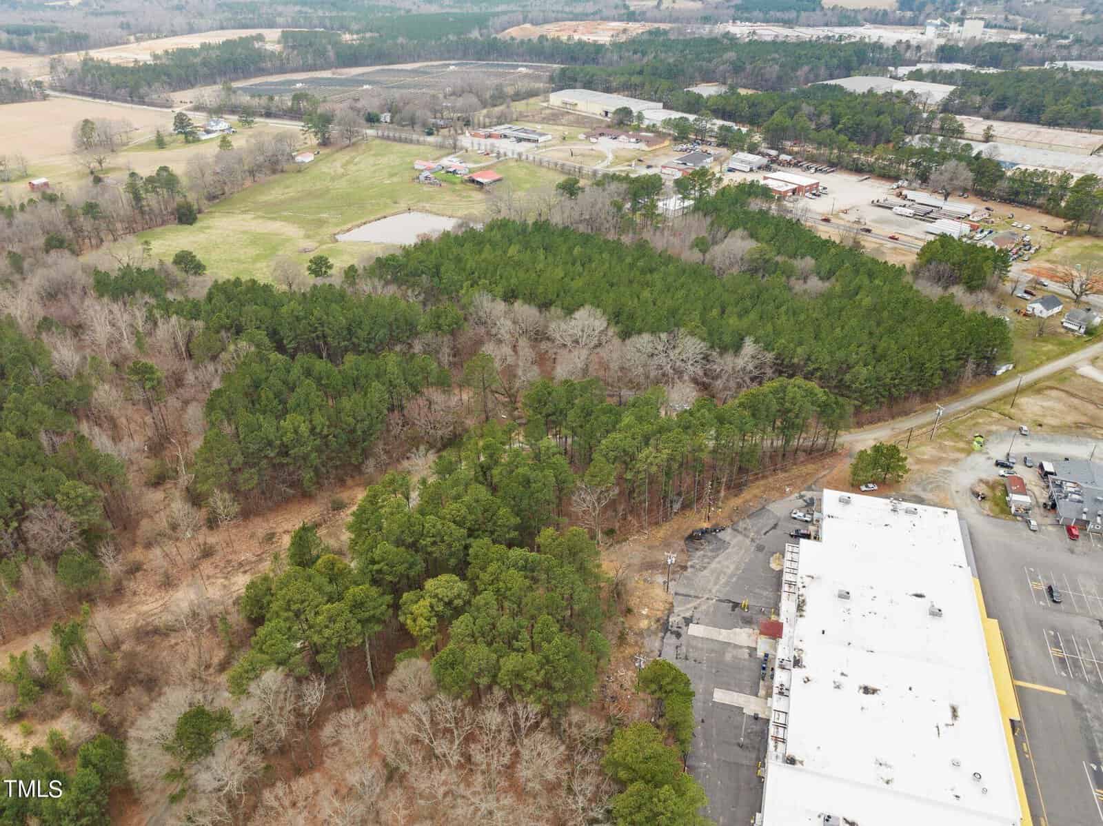 Aerial view of a large, wooded area surrounded by industrial buildings and a parking lot. The landscape features dense trees and open fields, with a few structures visible. A mix of green and brown vegetation can be seen across the terrain.