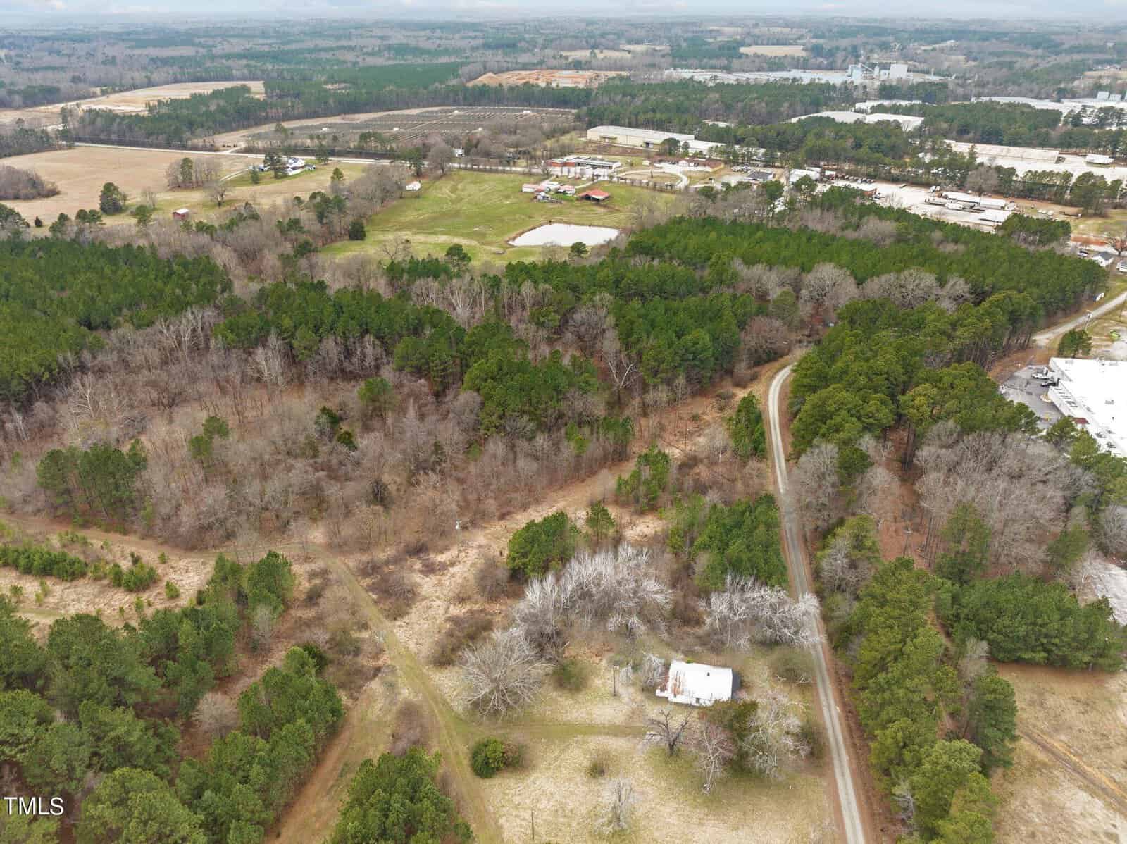 Aerial view of a rural landscape with dense forests, open fields, and a small road winding through. A few buildings are scattered throughout the greenery. A single house is visible near the bottom center of the image.