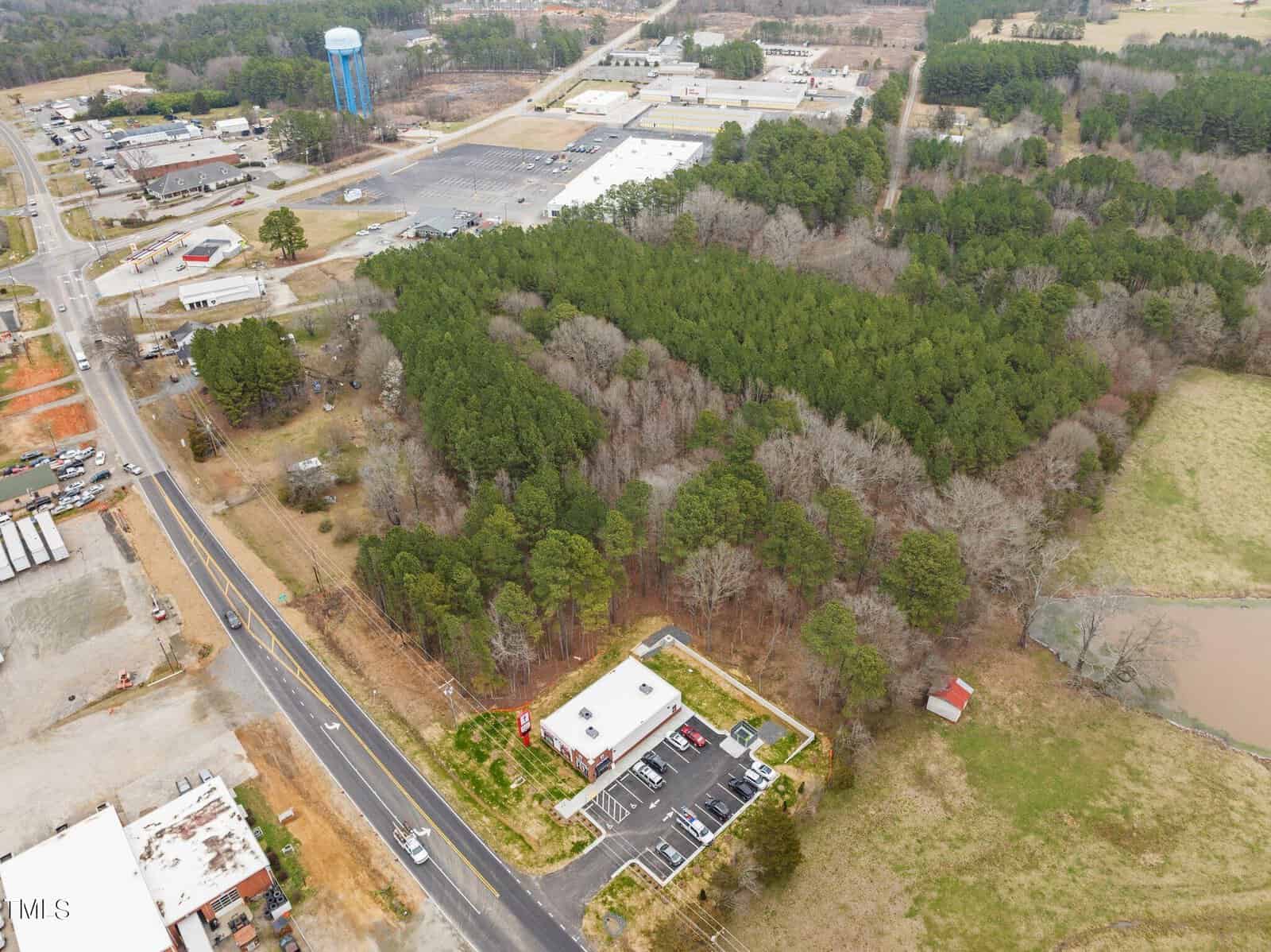 Aerial view of a rural area with a small commercial building and parking lot at the bottom. A large wooded area dominates the center. Roads and a few industrial buildings are visible nearby. A blue water tower stands in the upper left corner.