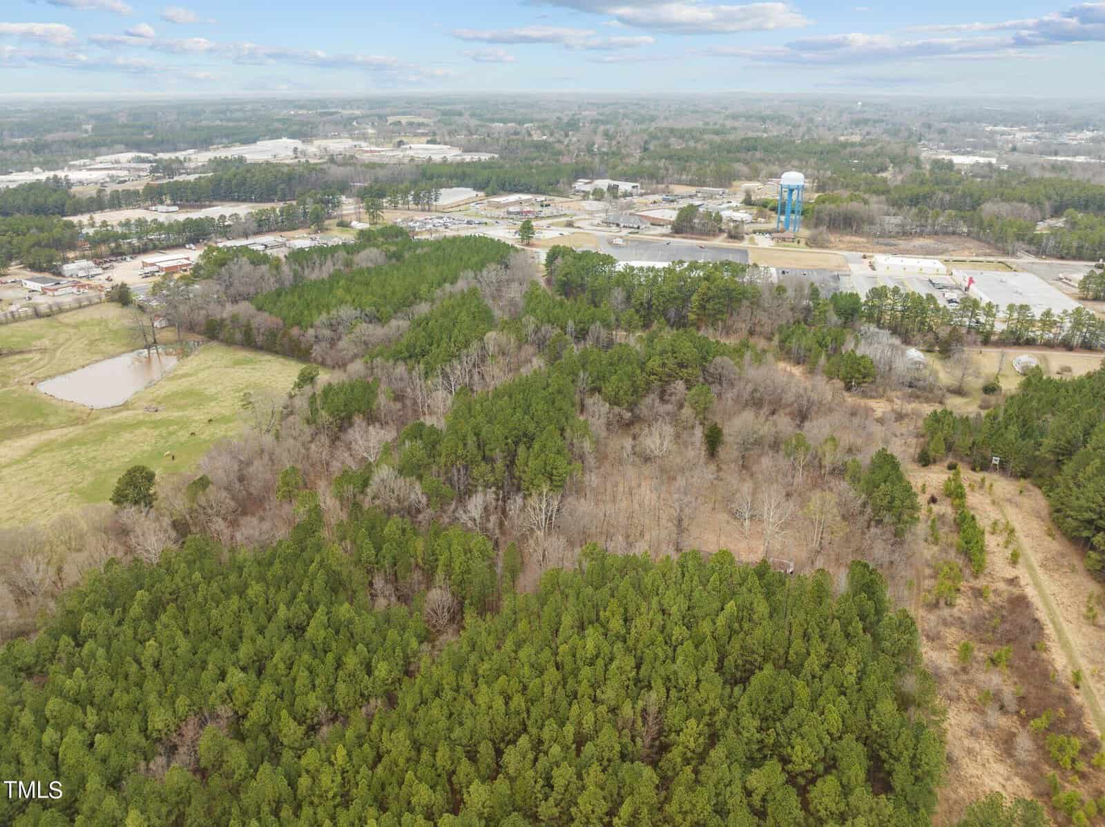 Aerial view of a rural landscape featuring a dense forest with a variety of trees. A small pond is visible on the left, surrounded by open fields. In the background, industrial buildings and a blue water tower can also be seen.