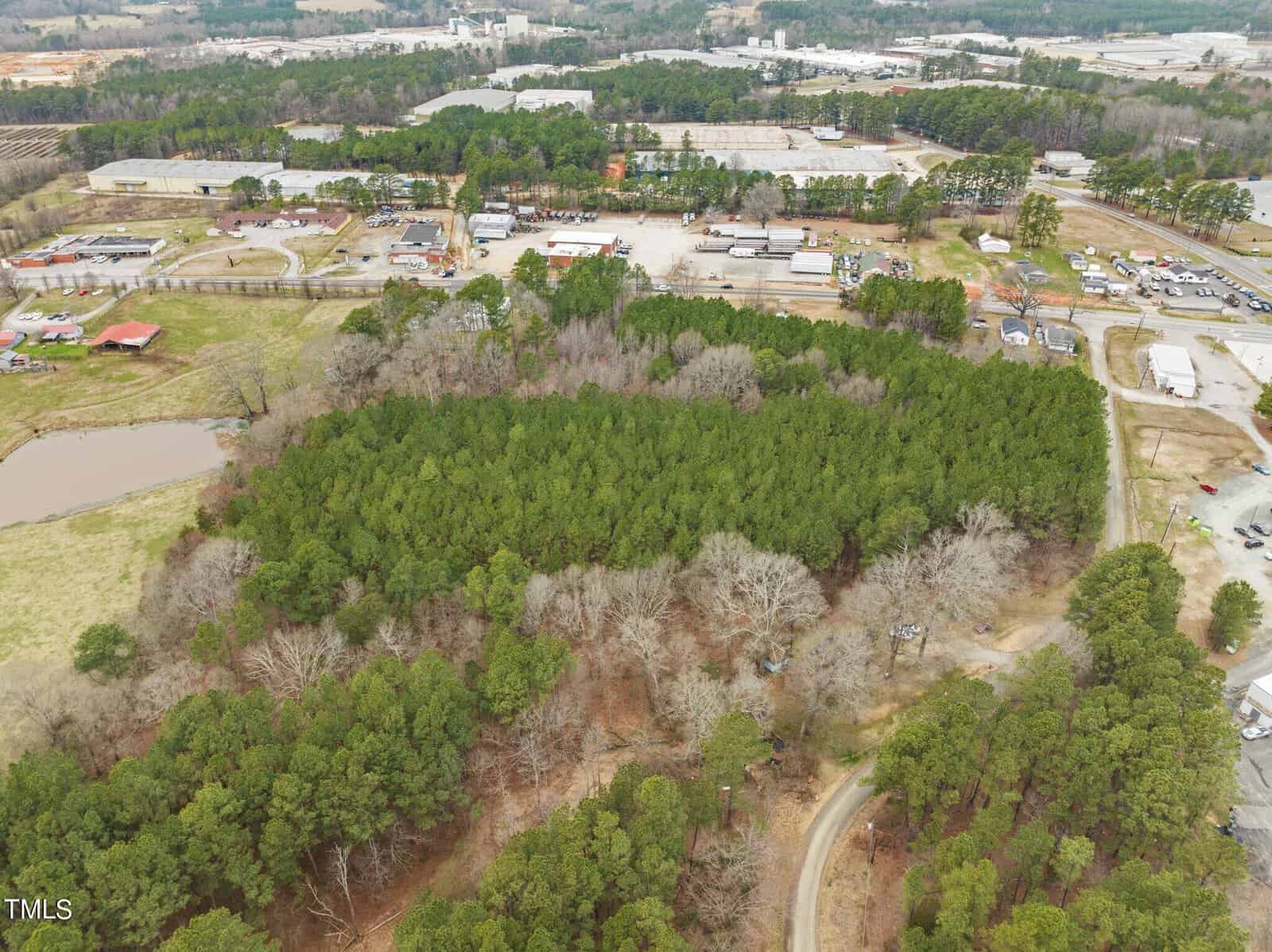 Aerial view of a lush, green forested area surrounded by rural roads and buildings. In the upper background, industrial structures and clearings are visible, with a pond and open fields on the left side of the image.