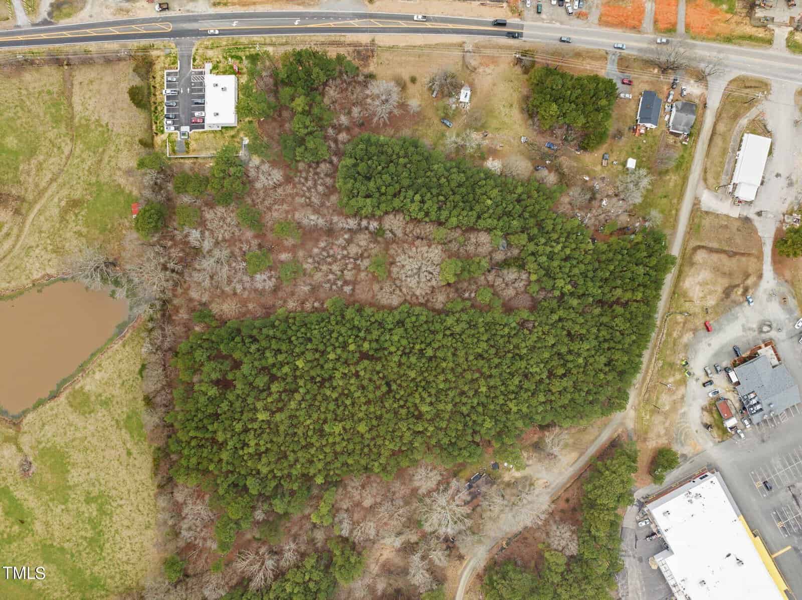 Aerial view of a wooded area with dense trees in the center, surrounded by roads and a few buildings. A small pond is visible in the bottom left corner. Roads border the top and right edges of the image.