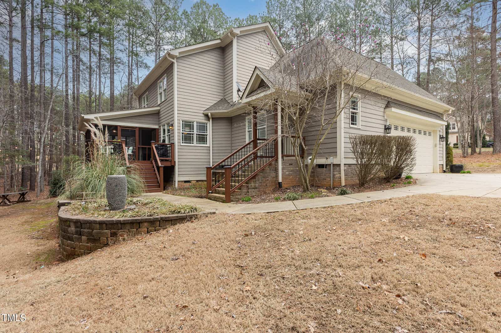 A two-story house with gray siding and a two-car garage is surrounded by tall trees. A wooden deck and stairs lead to the front entrance. The yard is dry, with a circular stone planter near the driveway. The sky is partly cloudy.