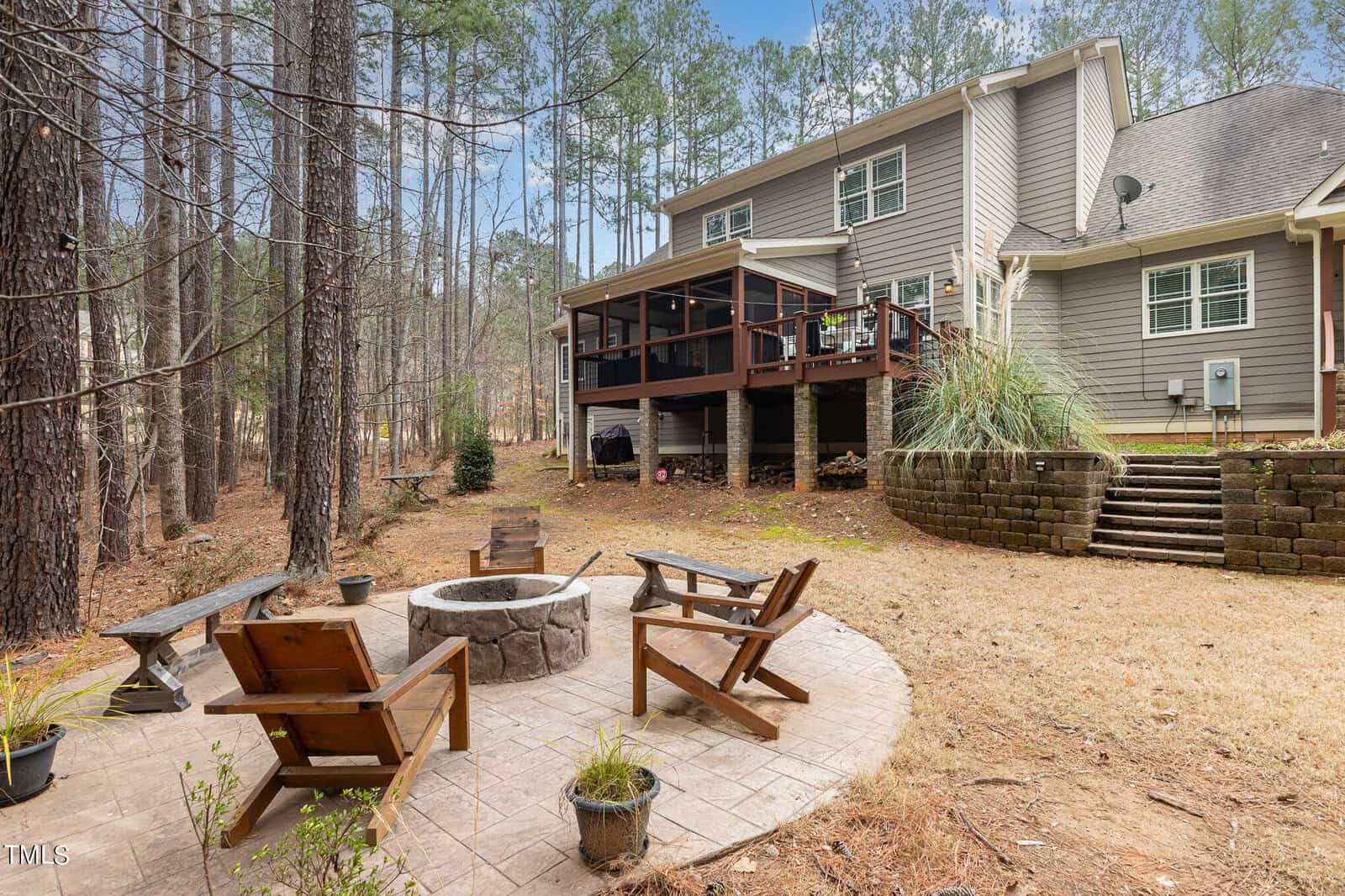 A backyard with a stone fire pit surrounded by four wooden chairs on a patio. Behind it, a multi-story house with an elevated deck and stairs. Tall trees line the background, creating a serene forest setting.
