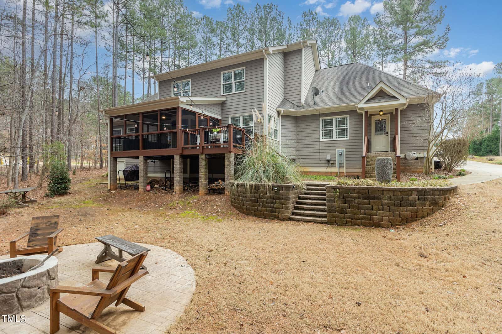 A suburban house with gray siding, surrounded by tall trees. The backyard features a raised wooden deck with a screened porch, a grassy area, and a stone fire pit with chairs. A stone retaining wall lines part of the yard. The sky is partly cloudy.