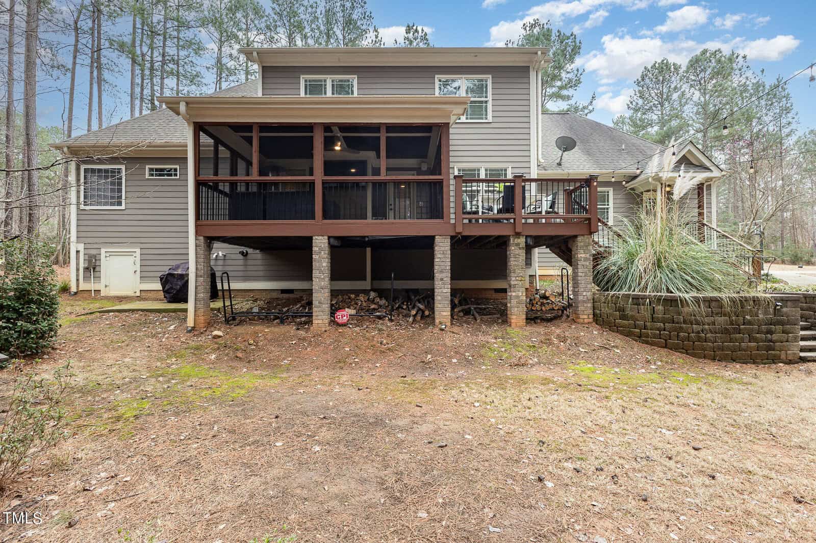 Rear view of a two-story house with a large wooden deck. The deck is elevated on stone pillars and overlooks a yard with pine trees in the background. A satellite dish is mounted on the side of the house. Stones line a small terraced section near the driveway.