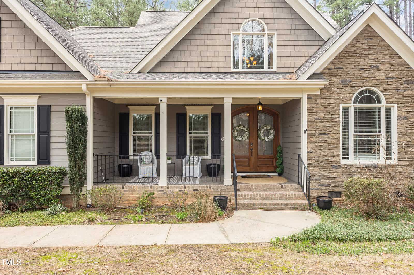A charming suburban house with a front porch featuring two rocking chairs and a pair of wreaths on double wooden doors. The exterior includes a mix of stone and shingle siding, black shutters, and a small upper window. A walkway leads to the entrance.