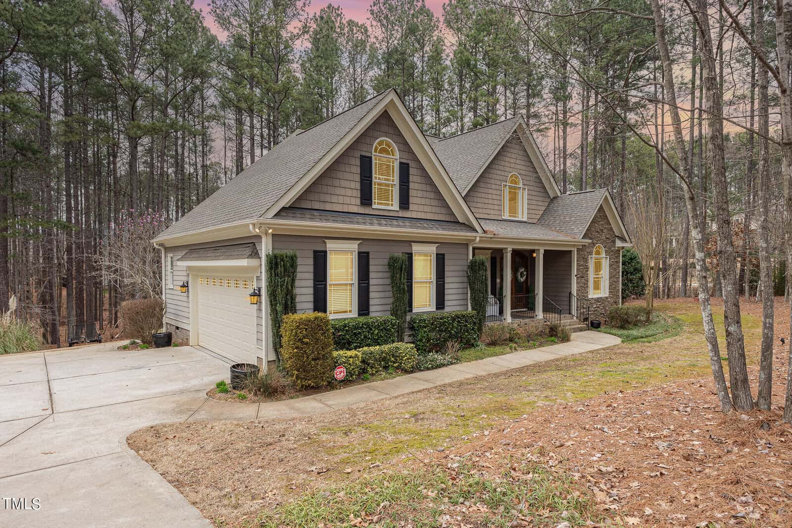 A charming two-story house with a front porch, surrounded by tall pine trees. The exterior is brown with white accents and a well-maintained lawn. A paved driveway leads to an attached garage. The sky is a soft pink at dusk.