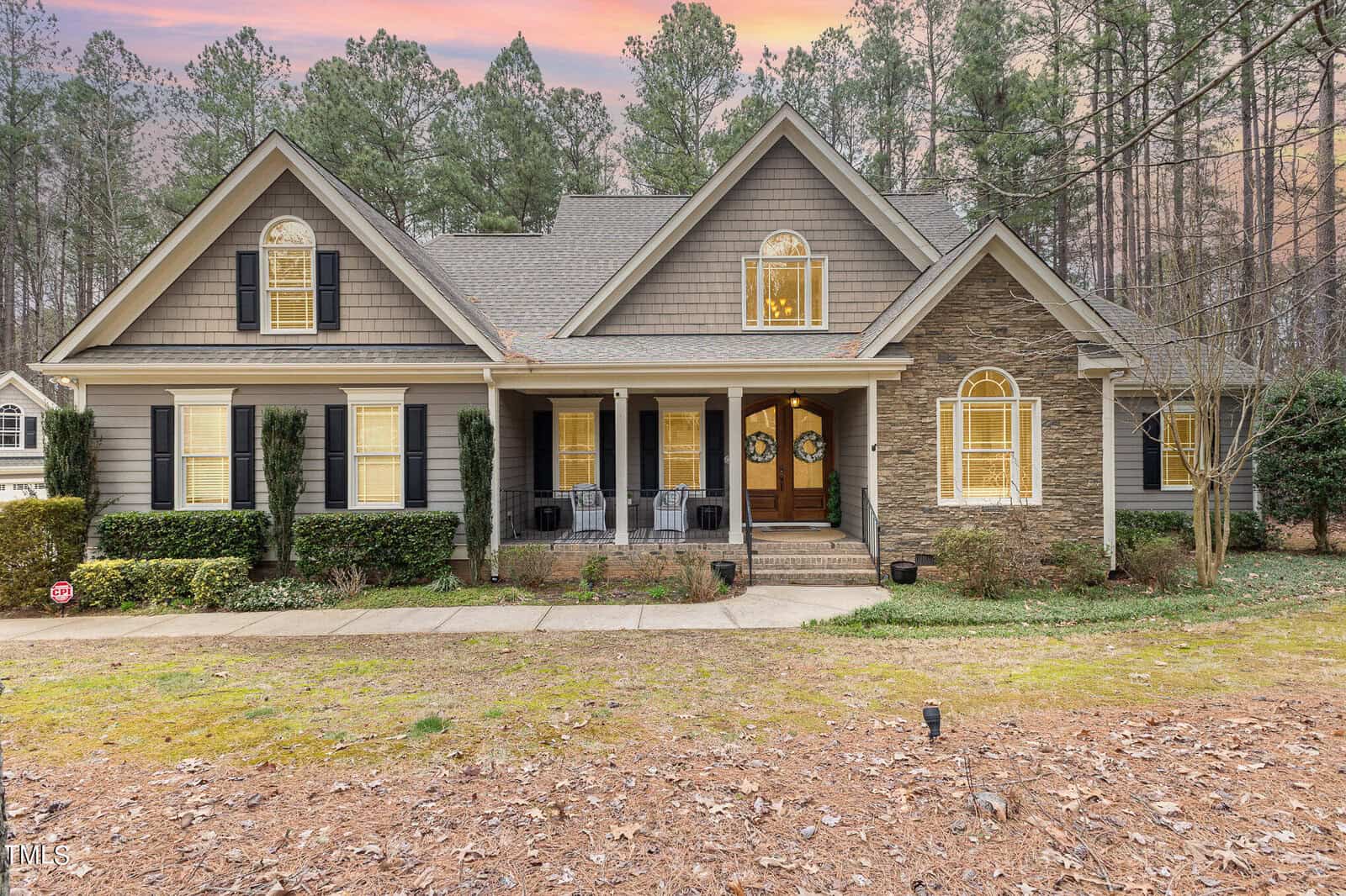 A large, elegant house with a shingle and stone facade, surrounded by tall trees. It features a front porch with black chairs, glowing windows, and a sunset sky in the background. A concrete walkway leads to the entrance.