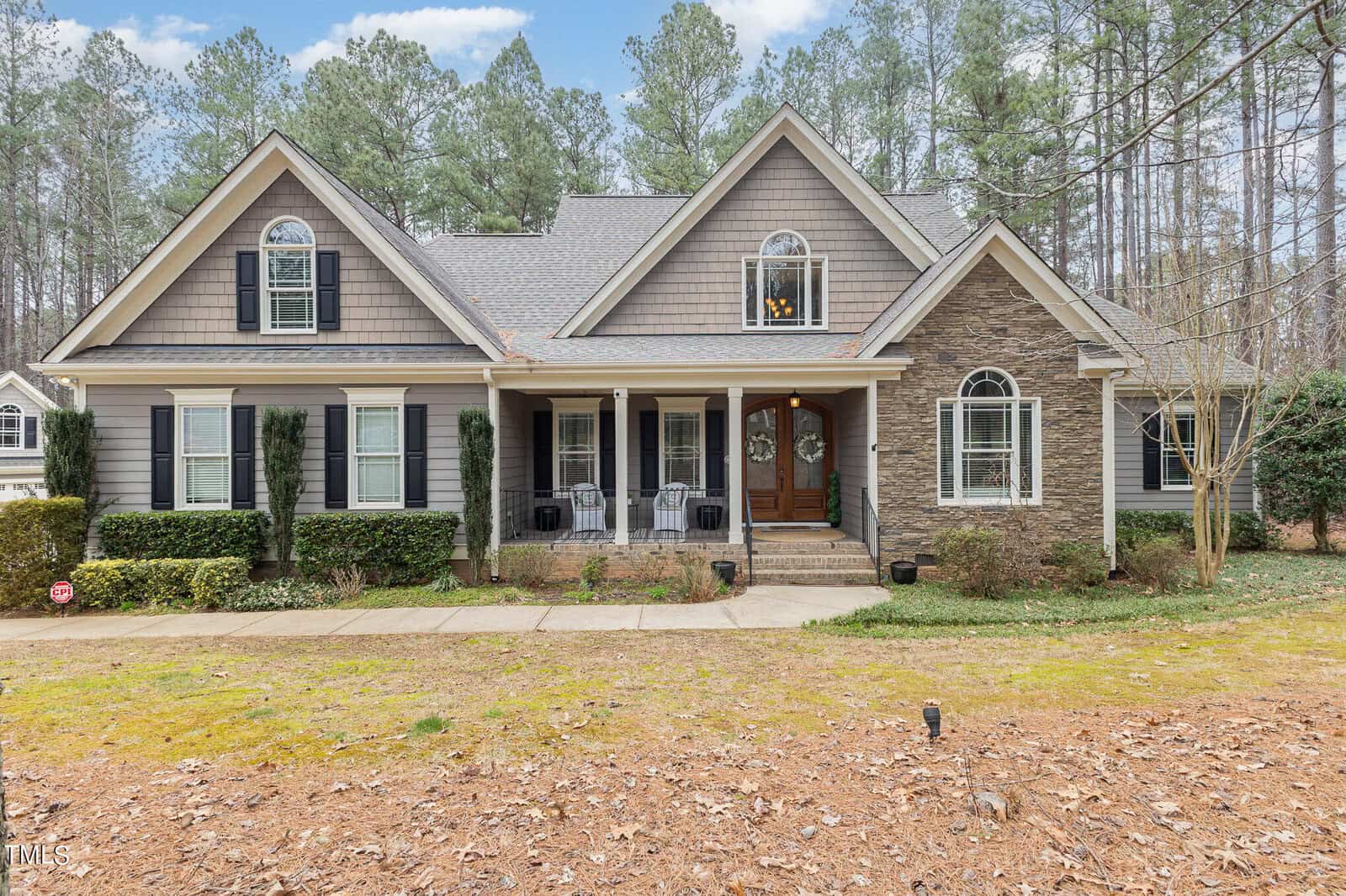A charming two-story house on 1199 Old Still Way, with gray siding and stone accents, surrounded by tall pine trees in Wake Forest. The front porch features white columns and black wicker chairs. The well-maintained lawn and shrubs add a welcoming touch.