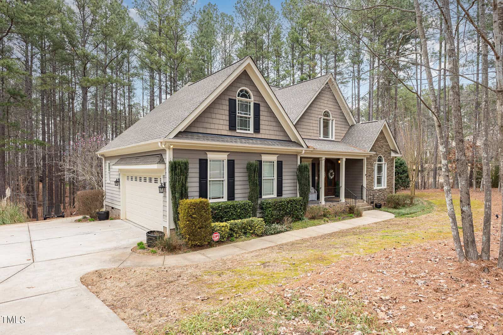 A suburban house with gray siding and a gable roof, surrounded by tall pine trees. The front has a two-car garage, white trim, dark shutters, and landscaped bushes. A concrete driveway leads up to the entrance, and the lawn shows some dry patches.