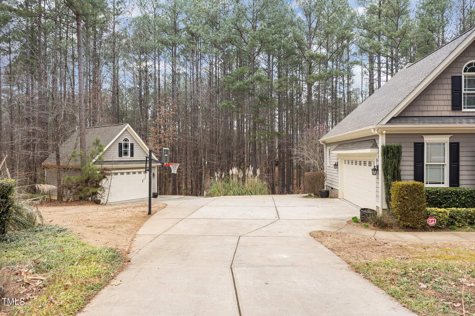 A driveway leads to a house and a detached garage, both with gray siding and white trim. A basketball hoop is positioned near the garage. In the background, a dense forest of tall pine trees is visible. The area is surrounded by manicured lawns.