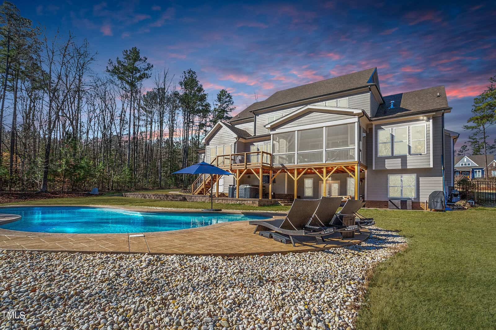 A two-story house with a large deck overlooks a backyard featuring a round swimming pool surrounded by a stone patio. Three lounge chairs are positioned by the pool. Trees and a neighboring house are in the background, under a colorful, cloudy sky.