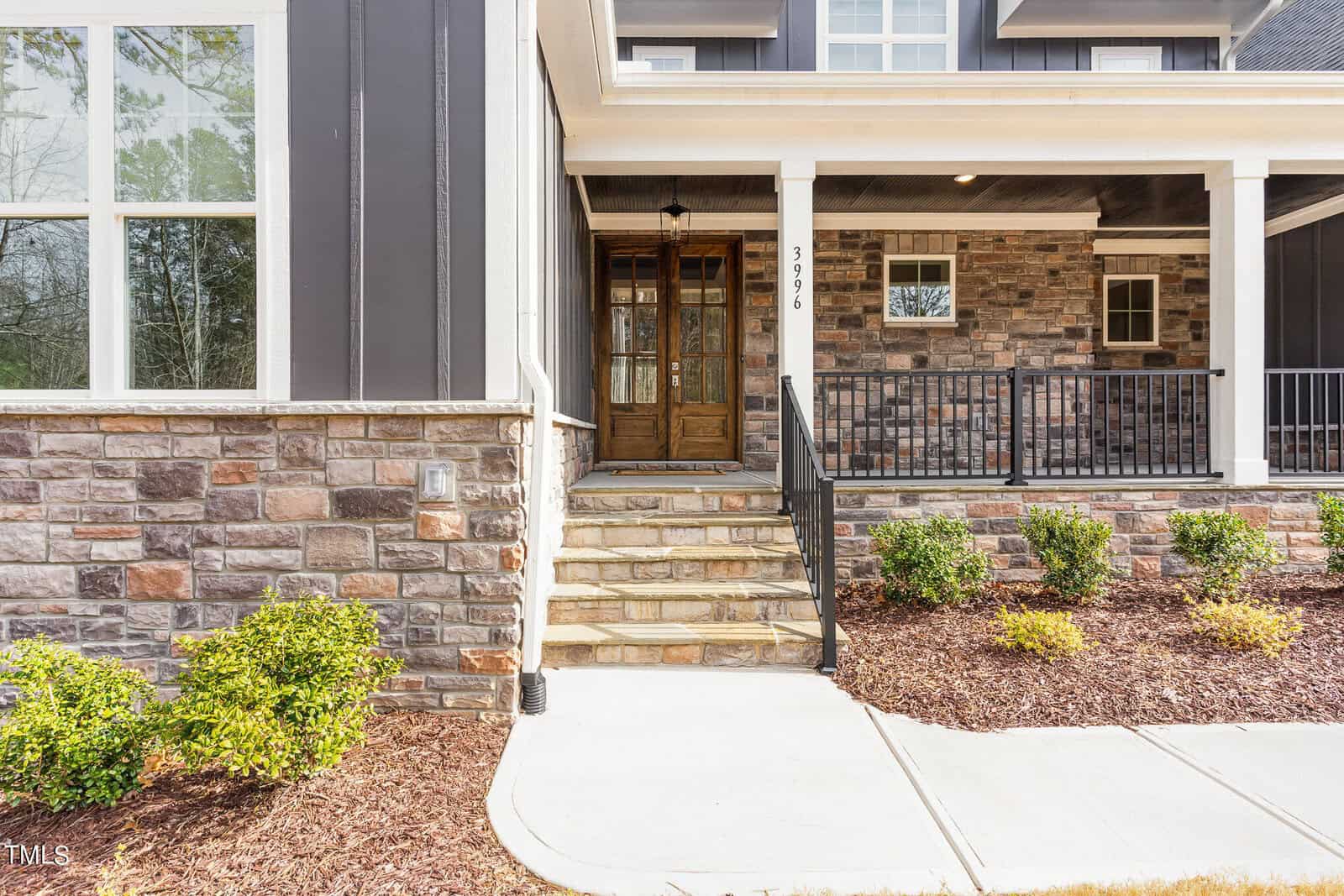 Front view of a modern house with stone and dark wood siding. It features a set of steps leading to double wooden doors, a small porch with railings, and landscaped bushes along the walkway.