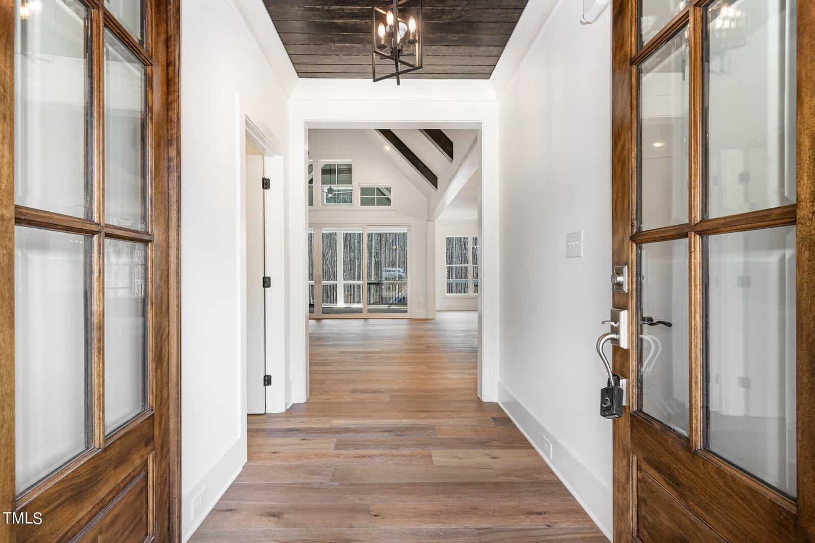 Open double doors reveal a spacious, modern hallway with wooden floors leading to a bright living area. High ceilings and large windows enhance the space, allowing natural light to flow in. Dark wood accents contrast with white walls and ceilings.