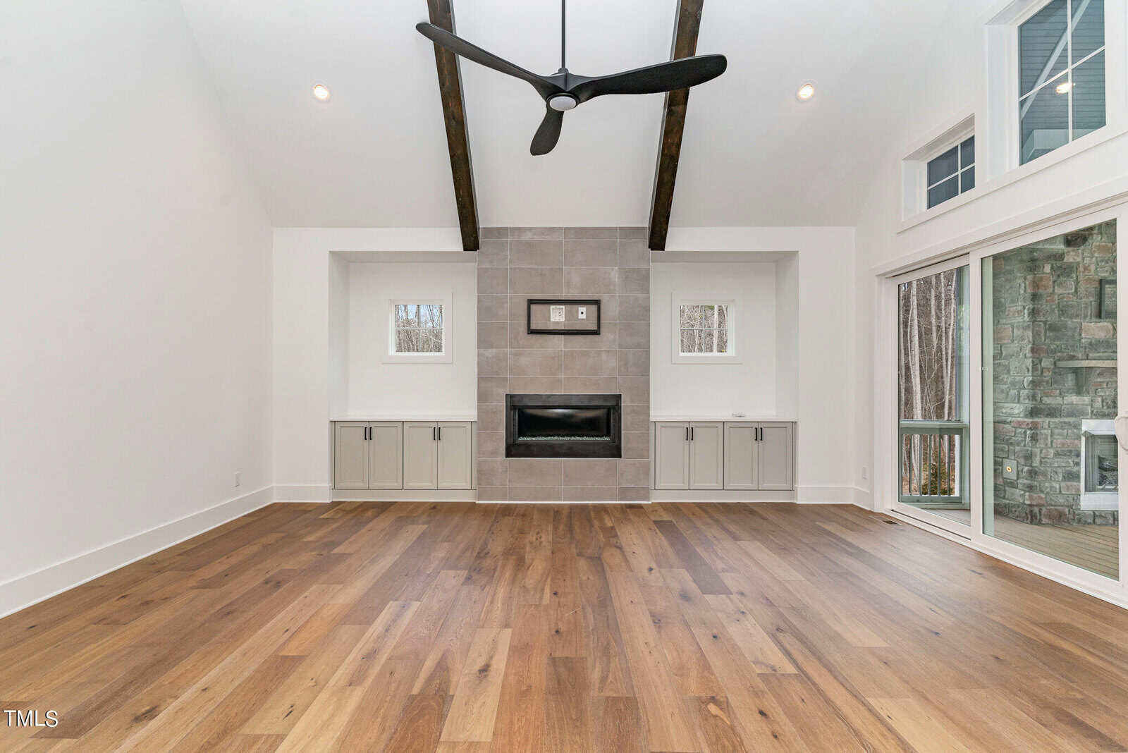 Spacious living room with a high ceiling, featuring a modern black ceiling fan, wood flooring, a central fireplace with tile surround, built-in cabinets on either side, and large windows with sliding glass doors.