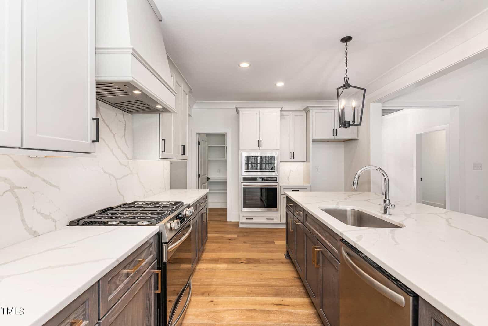 A modern kitchen with white cabinets, a marble-patterned backsplash, and a wooden floor. It features stainless steel appliances, including two ovens and a stove. A large island with a sink and pendant light completes the space.