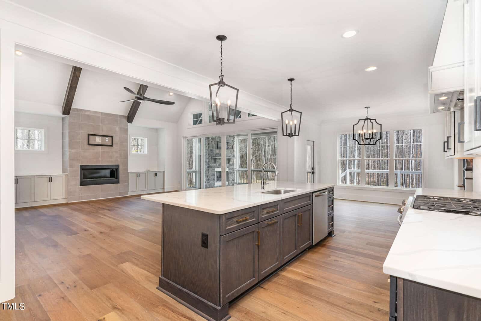 Modern open-concept kitchen and living space with hardwood floors, white walls, and large windows. The kitchen features a central island with a sink and pendant lights. The living area has a fireplace and exposed beam ceiling.