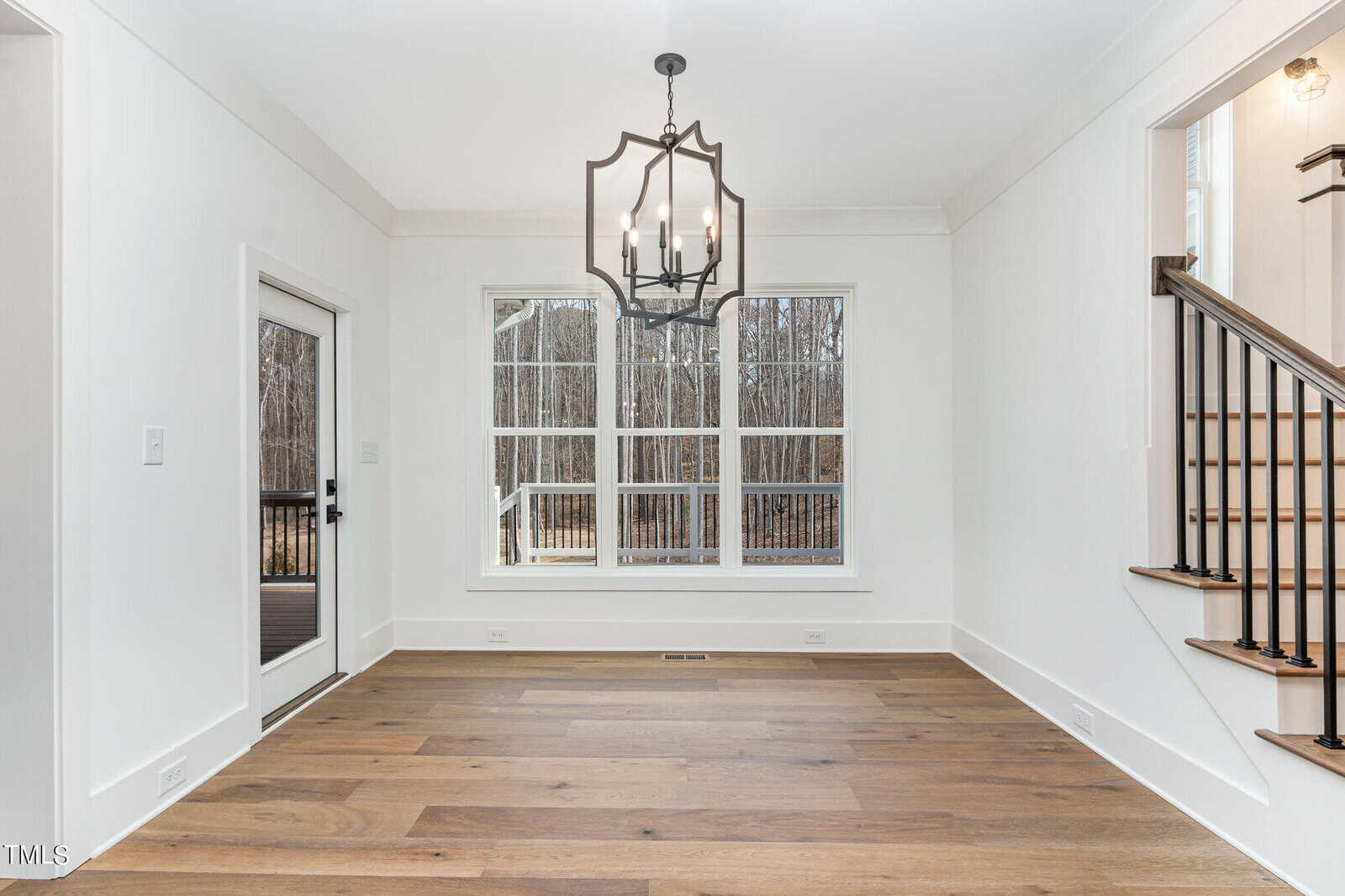 A bright, empty dining room features hardwood floors and a large window with a view of bare trees outside. A black chandelier hangs from the ceiling, and a sliding door to the left opens to a deck. A staircase is partially visible on the right.