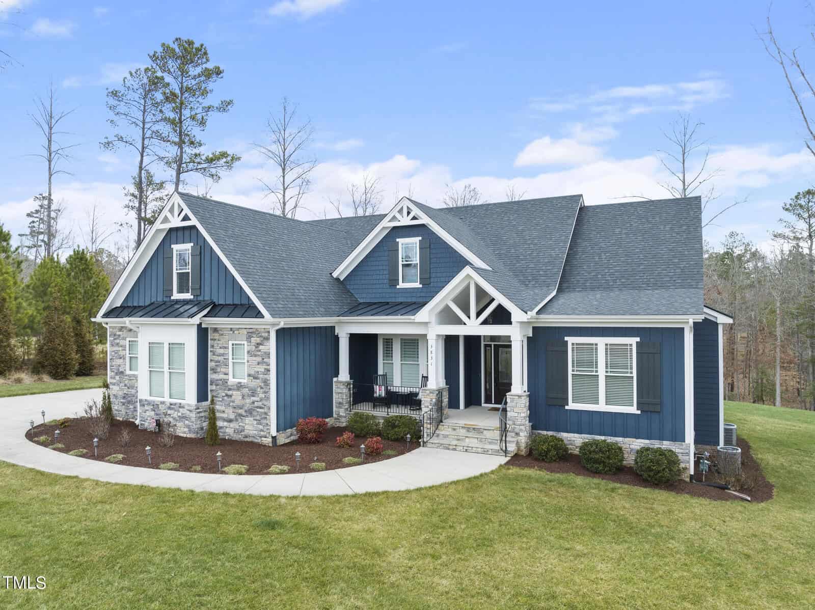 A modern, single-story house with blue siding and stone accents, featuring a large front porch and multiple gables. It's surrounded by a neatly landscaped yard with shrubs and a circular driveway, set against a backdrop of trees and a clear sky.