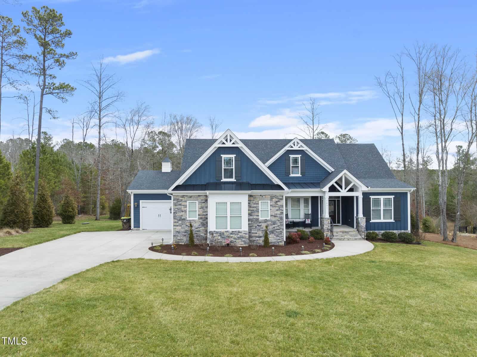 Located at 3831 Pickett Court in Wake Forest, this single-story modern house boasts a gray and blue exterior with white trims and stone accents. It features a two-car garage and large driveway, all surrounded by a well-manicured lawn and tall trees under a clear blue sky.