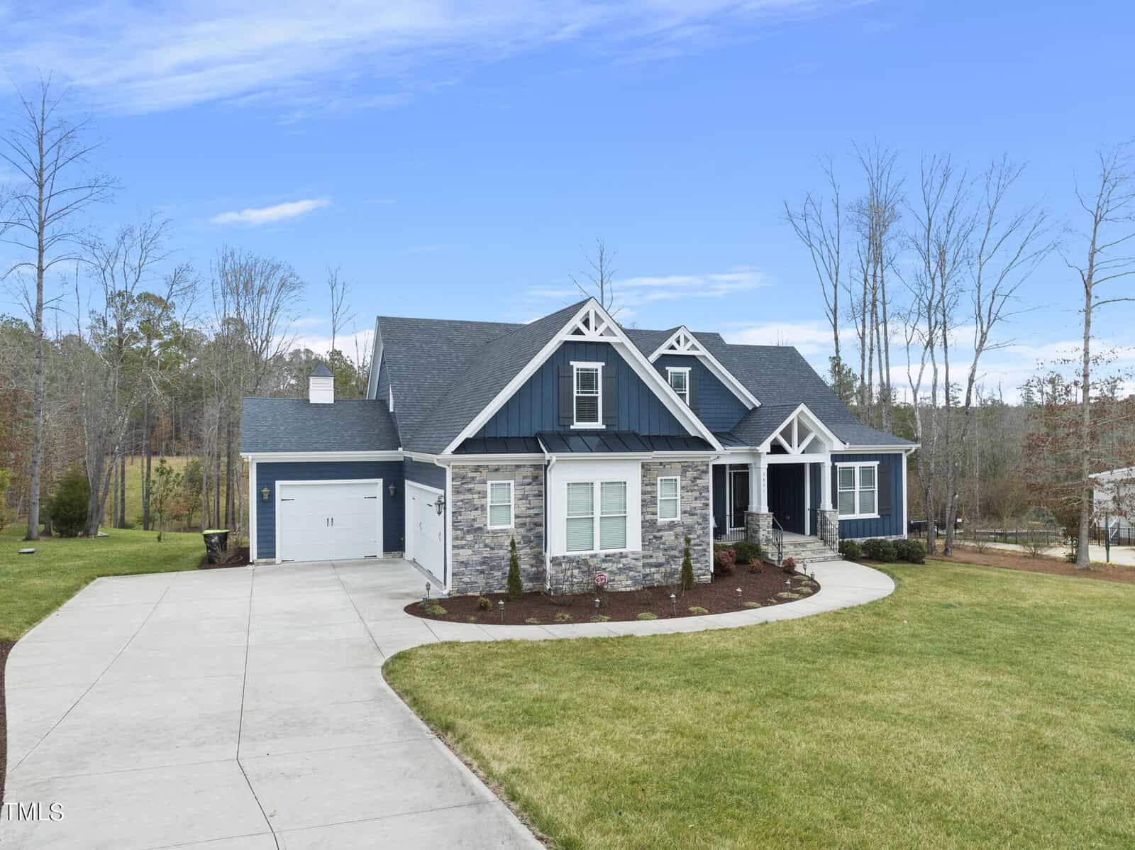 A modern, single-story house with stone and dark blue siding, featuring white trim and dormer windows. The home has a two-car garage and landscaped front yard with a driveway, surrounded by trees under a clear blue sky.