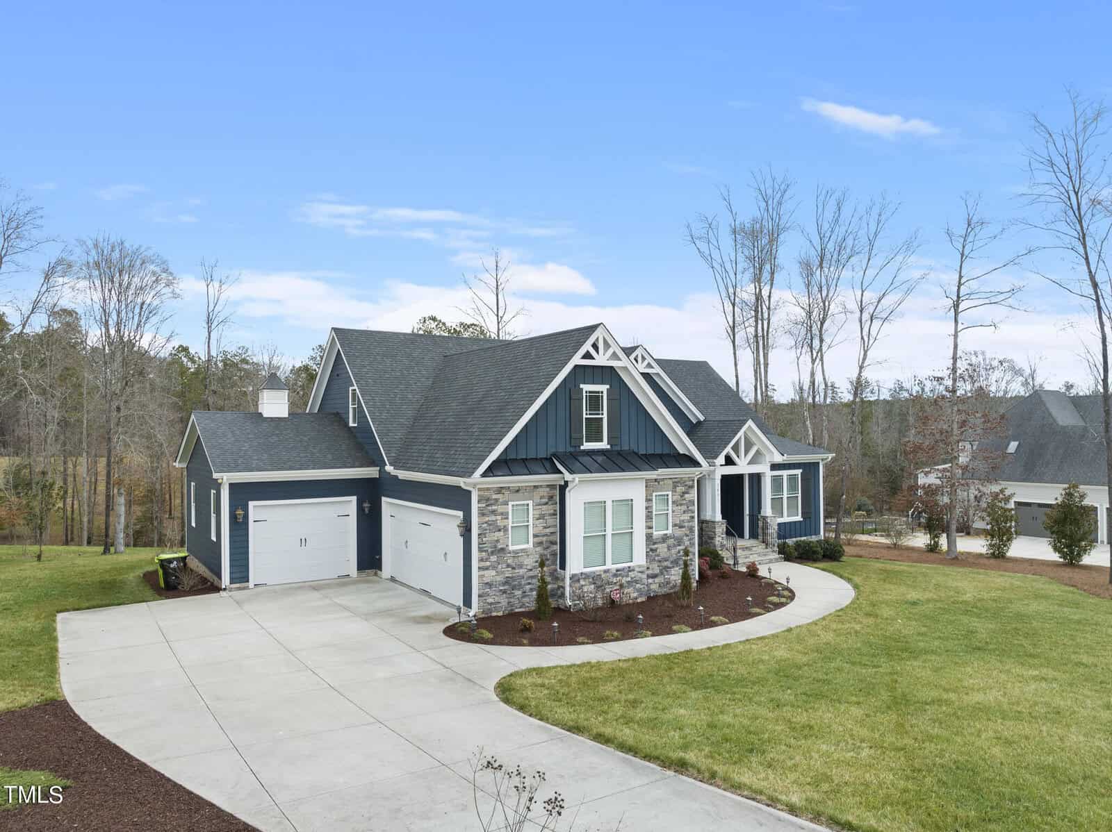 A charming suburban house with a stone facade and navy blue siding sits on a neatly landscaped yard. The house features a two-car garage and a curved driveway, surrounded by sparse trees against a clear blue sky.