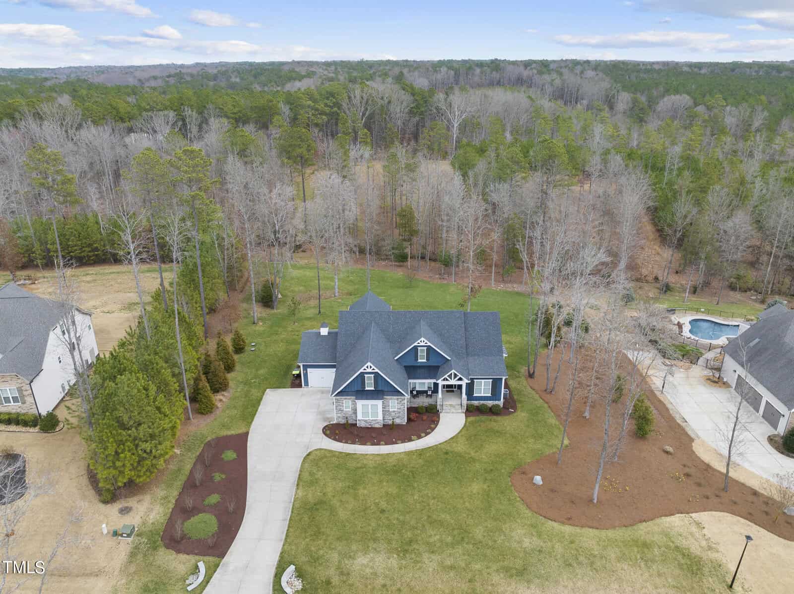 Aerial view of a large, single-story house with a dark roof and a landscaped front yard. The property includes a long driveway and is surrounded by trees. Nearby, a pool can be seen in the neighboring yard. Sparse forest extends in the background.