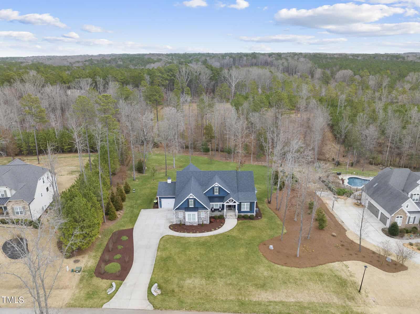 Aerial view of a suburban neighborhood with a focus on a large blue house. The house features a manicured lawn and a driveway. It's surrounded by trees and similar homes, with a clear blue sky above.