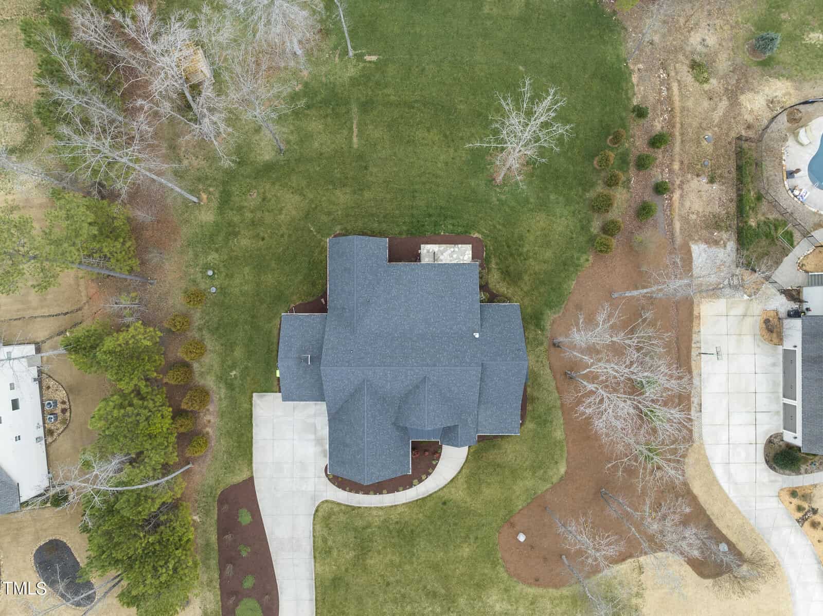 Aerial view of a house with a dark roof and surrounding yard. The property features a curved driveway, bare trees, and landscaped garden areas. Adjacent properties with similar features are visible. The grass is patchy in some areas.